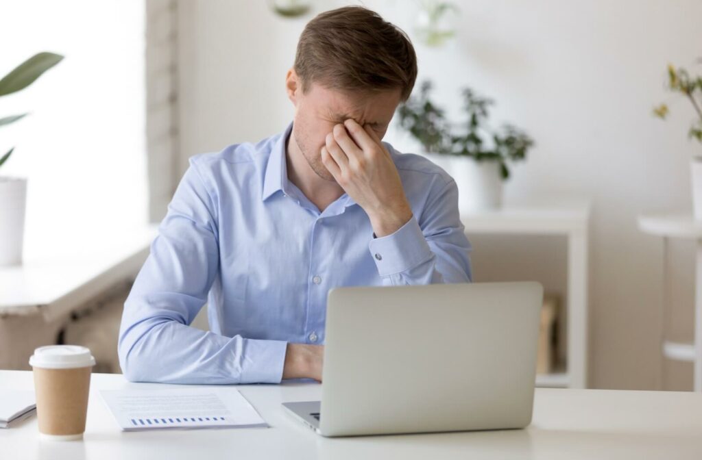 A man rubbing his eyes with his right hand while sitting in front of a laptop.