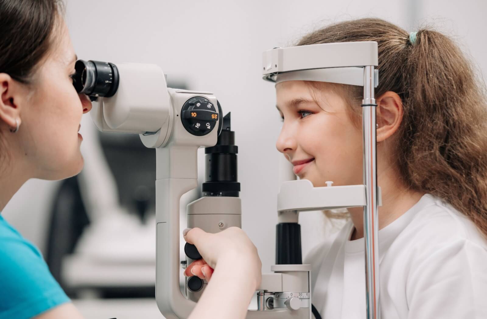 A child smiling while undergoing an eye exam with a slit lamp, assisted by an eye care professional.