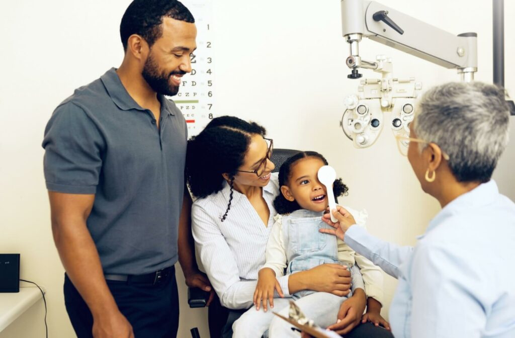 A child sitting on a parent's lap during an eye exam, smiling as the optometrist holds an occluder over one eye, with another parent standing nearby.