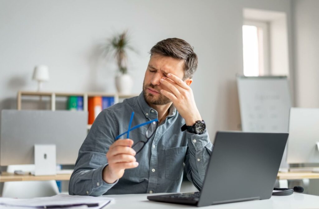 An individual sitting at a desk rubbing their eyes and holding eyeglasses while working on a laptop.