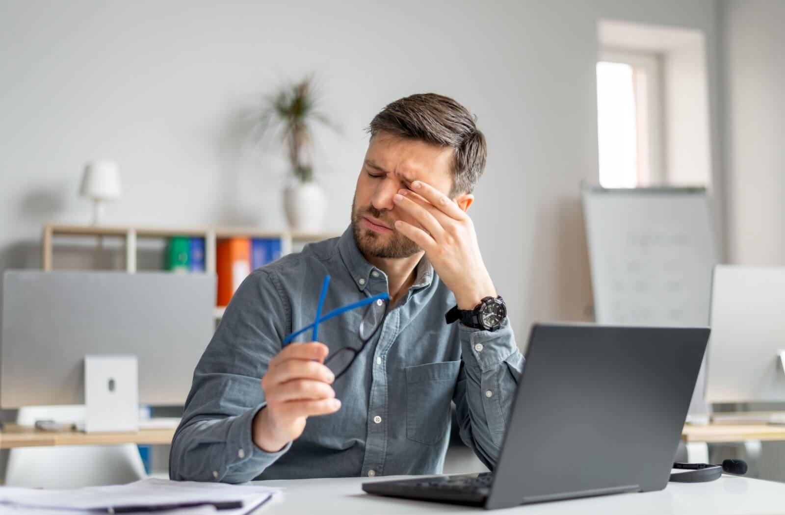 An individual sitting at a desk rubbing their eyes and holding eyeglasses while working on a laptop.