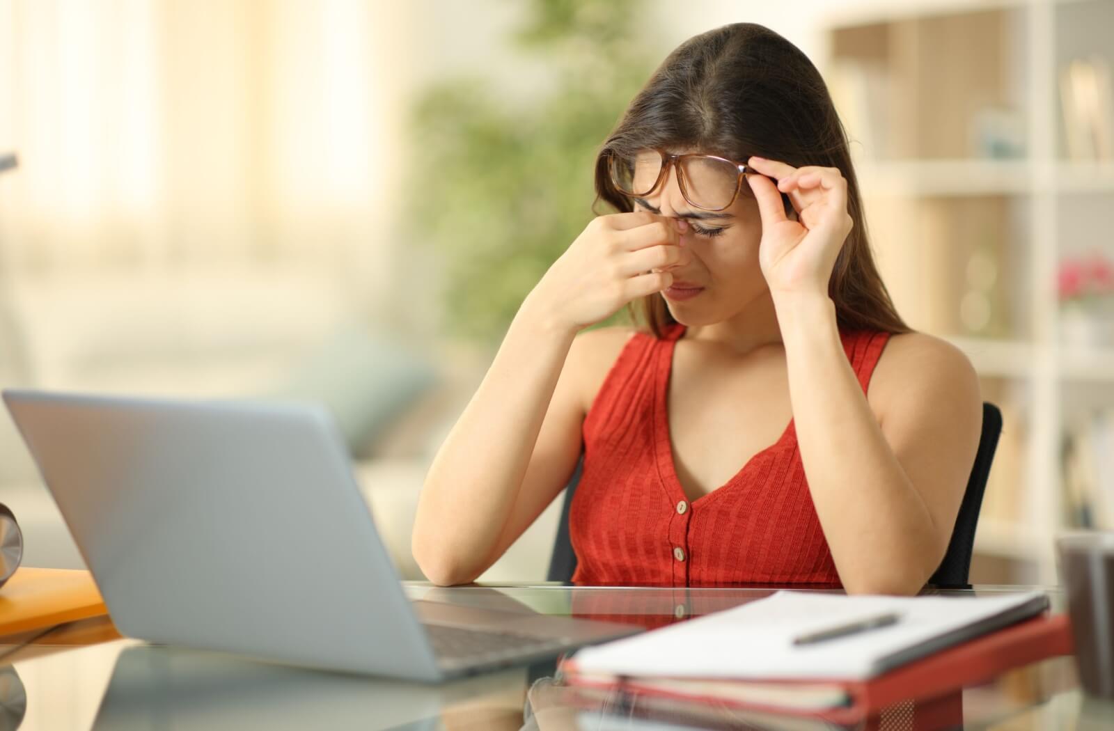 An individual lifting their glasses to rub the bridge of their nose while working at a laptop.